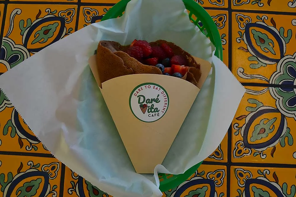 A vibrant, allergen-friendly crepe from Daré Vita Café filled with fresh berries, wrapped in branded paper on a colorful, decorative tile background, showcasing their commitment to healthy, wholesome meals.| Daré Vita Café | Las Vegas
